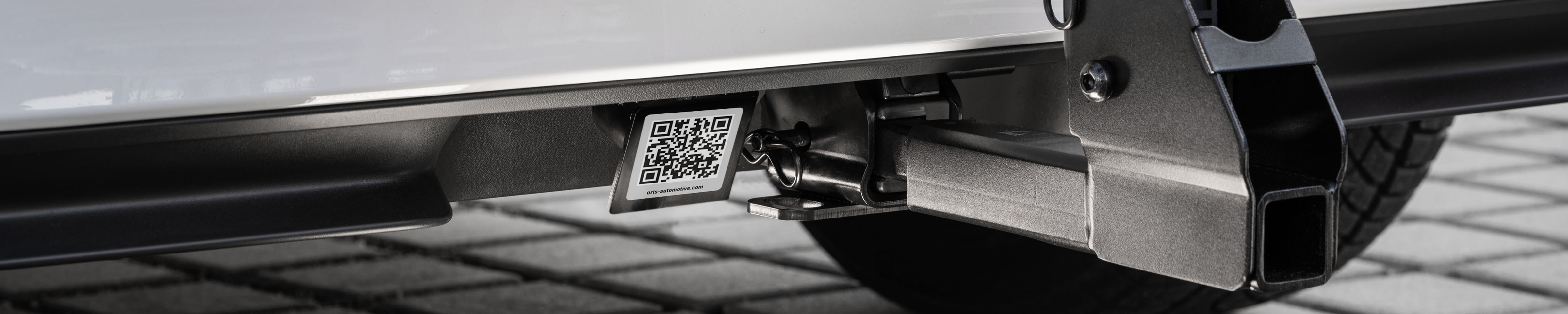Close-up of an ORIS trailer hitch receiver with integrated QR code label mounted beneath a vehicle. The image shows the square receiver opening and mounting point under the rear bumper.