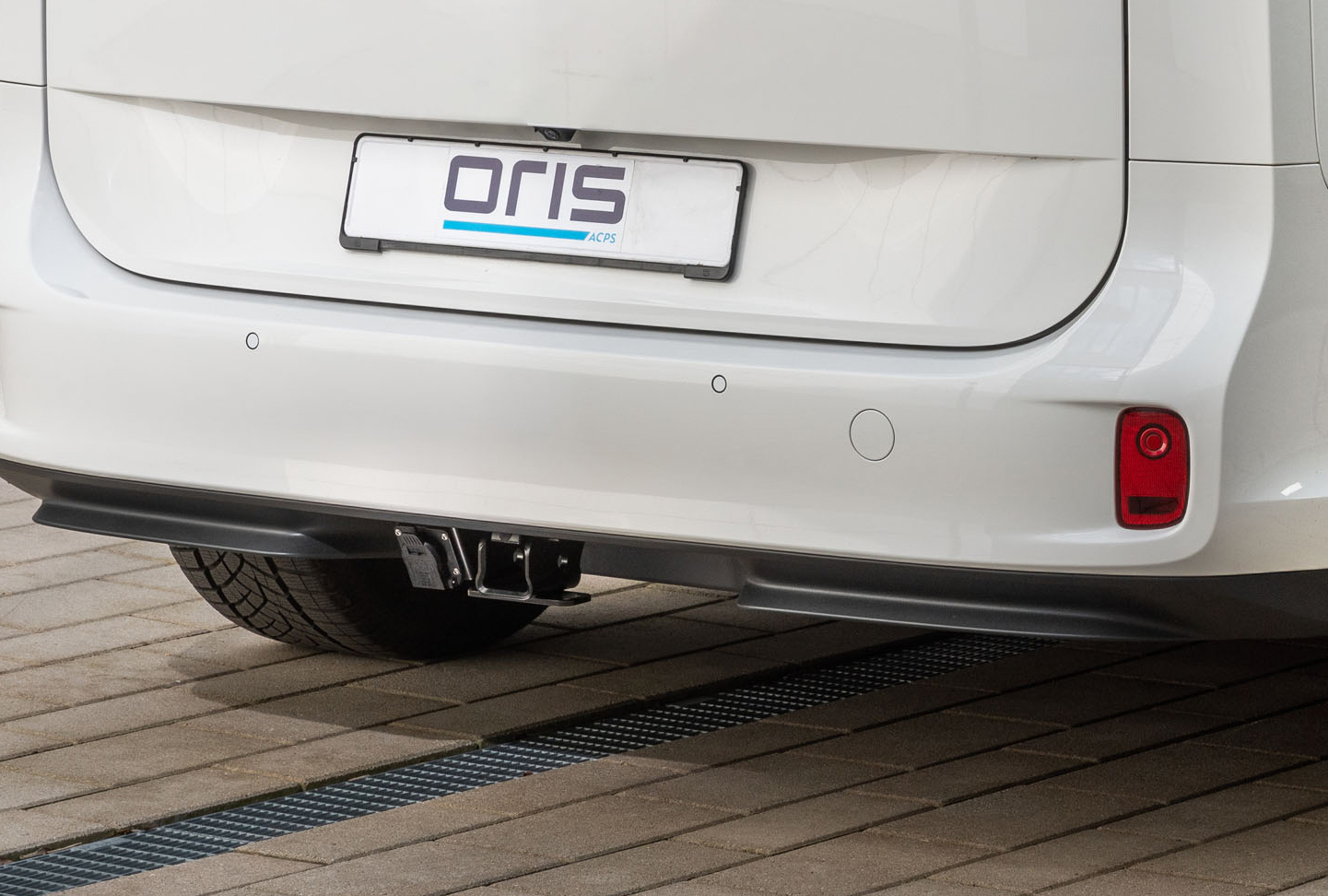 Rear view of a white vehicle with integrated ORIS receiver hitch and electrical socket mounted beneath the rear bumper on a paved surface.