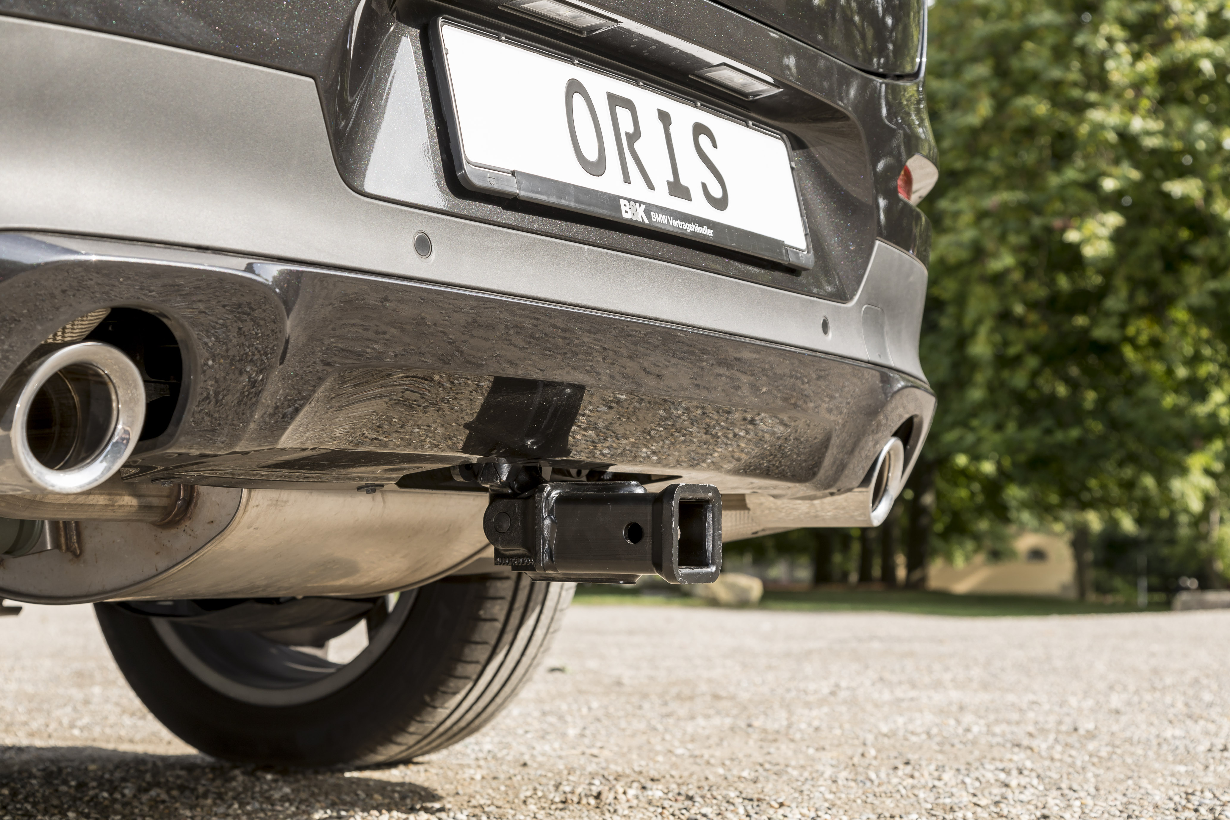 Rear view of a dark grey vehicle with ORIS detachable receiver hitch mounted beneath the rear bumper, shown on a gravel surface with trees in the background.